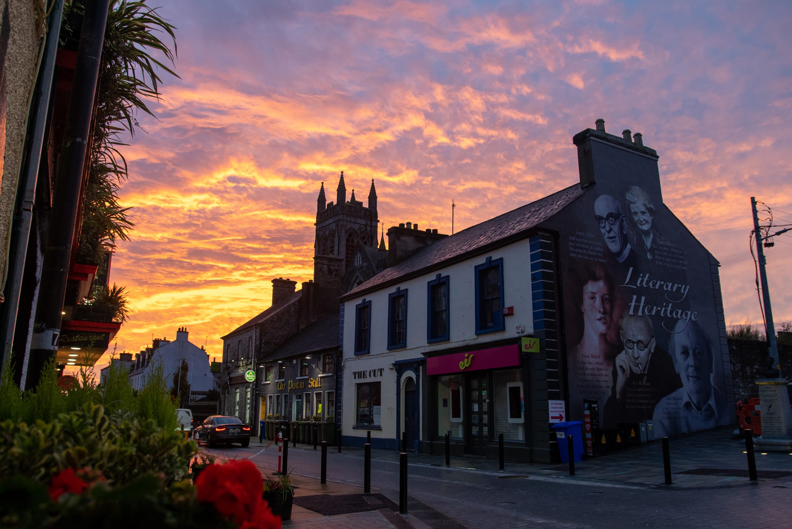 Street scene with a mural of literary figures on a building, framed by a dramatic sunset sky and a church tower in the background, from Ireland's Hidden Heartland.