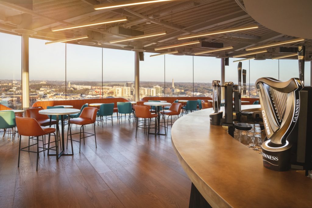 Interior of a modern bar with colorful seating and large windows offering a panoramic view of the city skyline, featuring Guinness taps on the counter.