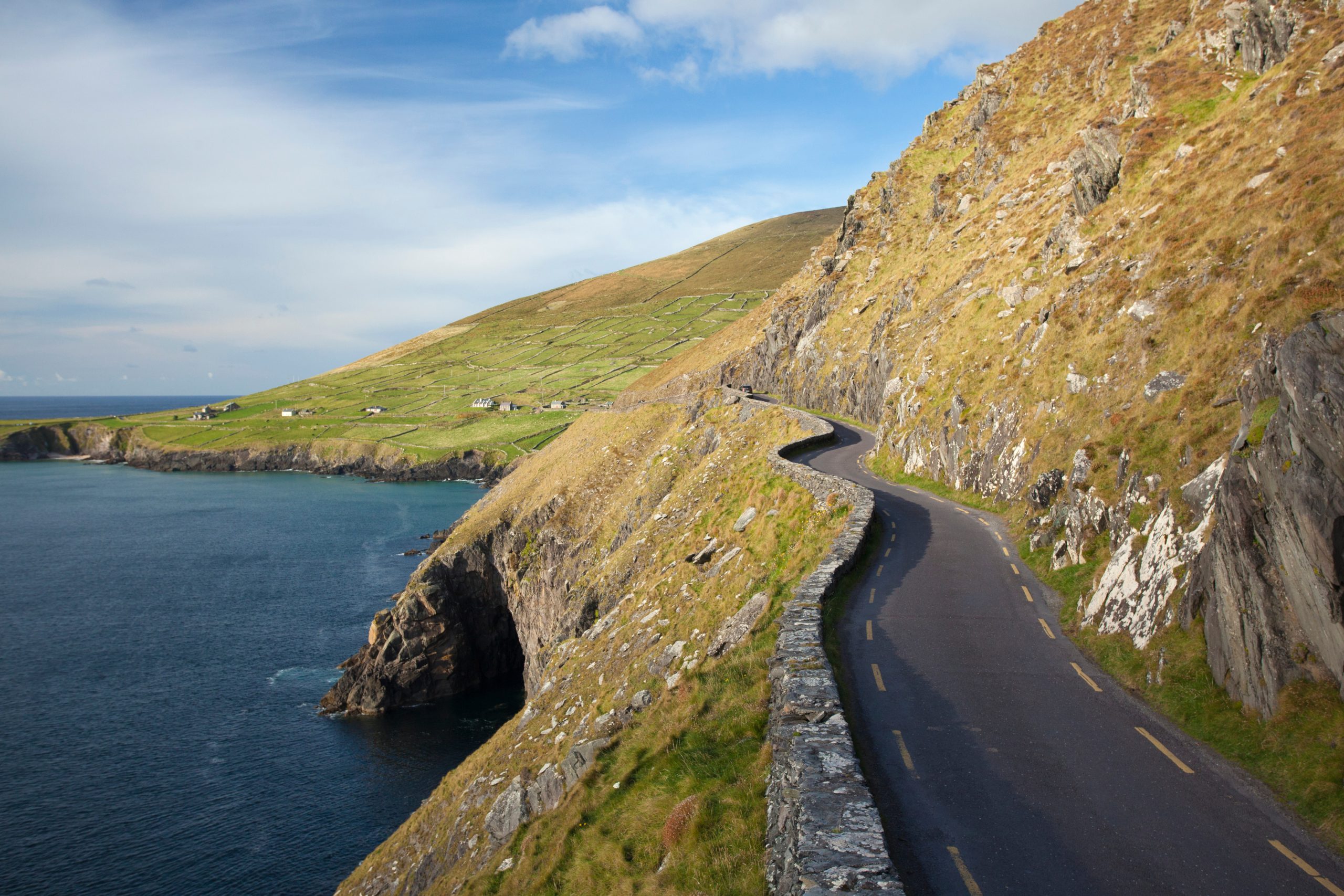 A narrow road winding along the steep cliffs of Slea Head, with the rugged coastline and rolling hills of the Dingle Peninsula in the background.